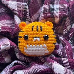 May include: A crocheted tiger head with orange and black stripes, black eyes, and white teeth. The handmade tiger is set against a plaid background of burgundy, white, and black.