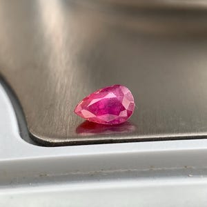Peut inclure: Une pierre précieuse à facettes en forme de poire, d'une couleur rose-rouge profonde et vibrante. La pierre présente de multiples facettes triangulaires et en forme de cerf-volant qui captent la lumière, créant des reflets internes.