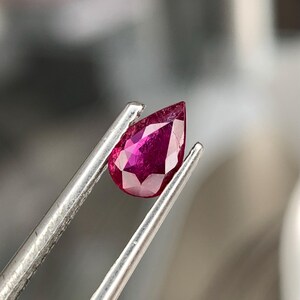 以下が含まれることがあります： 銀色のピンセットで保持された、梨型のファセットルビーの宝石。ルビーは深く鮮やかな赤色で、複数のファセットが光を捉えています。宝石は焦点であり、その透明度と輝きを示しています。