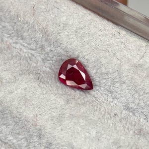 Puede incluir: Una gema de rubí facetada en forma de pera. La piedra roja oscura tiene una superficie pulida y se encuentra sobre un fondo gris suave. La gema está sujeta por una herramienta de metal, mostrando su corte y claridad.