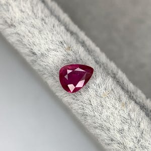 Op de afbeelding: Een peervormige, gefacetteerde robijn edelsteen. De robijn heeft een diepe, levendige rode kleur en ligt op een grijze, getextureerde achtergrond. De edelsteen heeft een gepolijst oppervlak dat licht reflecteert.