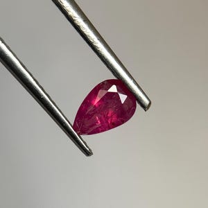 Könnte beinhalten: Ein facettierter, birnenförmiger Rubin-Edelstein, der von einer silbernen Pinzette gehalten wird. Der Rubin hat eine tiefe, lebendige rote Farbe mit sichtbaren inneren Einschlüssen. Die Pinzette ist schräg vor neutralem Hintergrund positioniert.