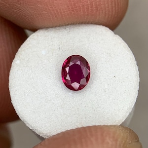 以下が含まれることがあります： 楕円形にカットされたルビーの宝石。深い赤色で、白い表面に展示されています。宝石のファセットは光を反射し、その輝きを示しています。クローズアップビューは、宝石の透明度と色の彩度を強調しています。