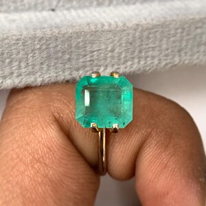 Puede incluir: Un anillo de oro con una gran esmeralda rectangular. La esmeralda es de un verde vibrante y está engastada en una fina banda de oro. El anillo se muestra en un dedo, sobre un fondo suave y neutro.