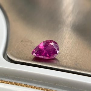 Puede incluir: Una gema facetada en forma de pera, de color rosa intenso, descansa sobre una balanza plateada. El corte de la gema revela múltiples facetas, realzando su brillo. El color de la piedra es un rosa vibrante y saturado.