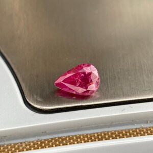 Peut inclure: Une pierre précieuse en forme de poire avec une teinte rose profonde et vibrante et une surface à facettes qui reflète la lumière. La pierre présente un sommet pointu et une base arrondie, mettant en valeur sa clarté et sa finition polie.