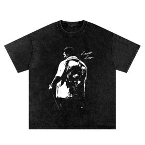 May include: Black t-shirt with a faded, vintage look. The shirt features a white graphic of a person from the back, wearing a jacket. The words "Lant Lay" are written in white script on the upper right side of the shirt.