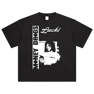 May include: Black t-shirt with a white graphic. The graphic includes the text "MONEY DR*GS" vertically on the left side, the name "Lucki" in cursive, and a black and white image of a person holding an object.