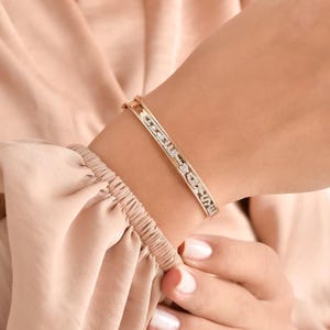 Puede incluir: Una pulsera dorada con la inscripción "Love Me Forever" en una fuente delicada, adornada con pequeñas piedras brillantes. La pulsera es de estilo articulado, diseñada para ser usada en la muñeca.