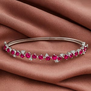 May include: A silver bracelet with a row of vibrant red gemstones and small white accent stones. The bracelet is set against a backdrop of a draped, muted rose-colored fabric.