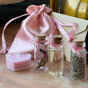 Könnte beinhalten: Eine Anordnung mit einer herzförmigen rosa Kerze, kleinen Glasflaschen mit Korken und einem rosa Satin-Kordelzugbeutel. Die Flaschen enthalten getrocknete Blumen, eine klare Flüssigkeit und getrockneten Lavendel. Ein kleiner Briefumschlag aus Kraftpapier mit rosa Band.