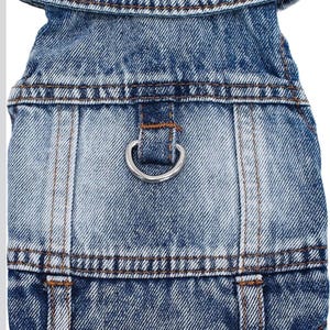 May include: A blue denim jacket featuring a folded collar, visible brown stitching, and a silver-toned metal D-ring attached to the center back panel. The garment has a classic acid-wash texture with vertical seams and a structured hemline.
