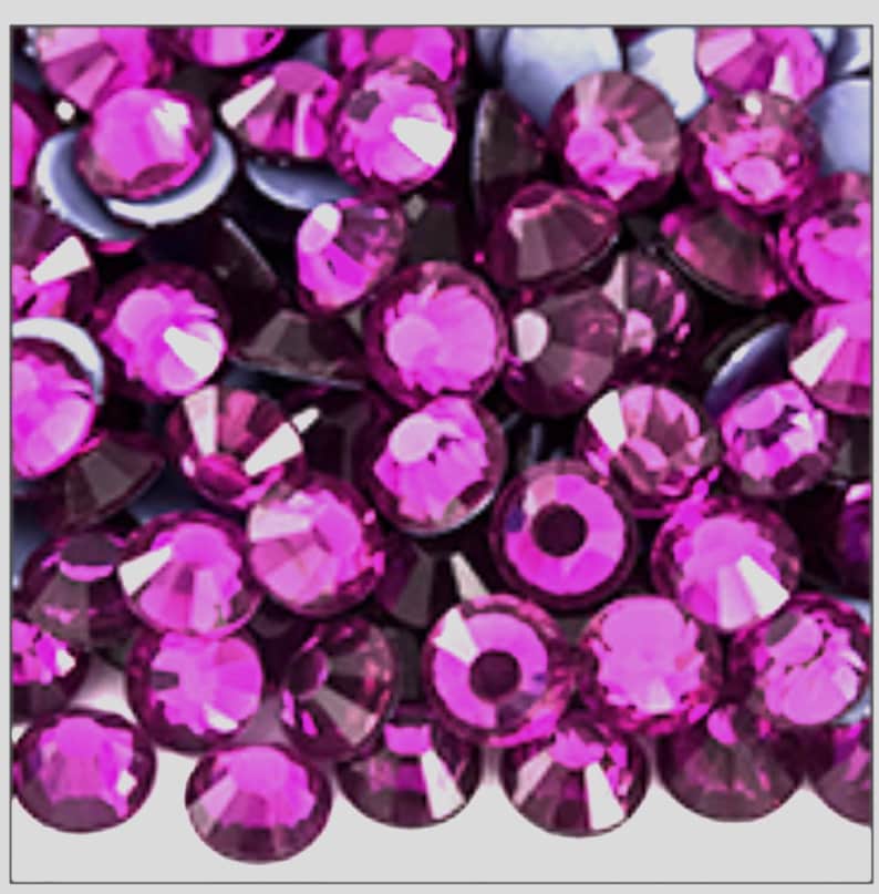 May include: A collection of numerous small, round, faceted magenta rhinestones. Each individual stone features a brilliant cut with multiple reflective surfaces, creating a vibrant, deep pink appearance across the entire group of loose gems.