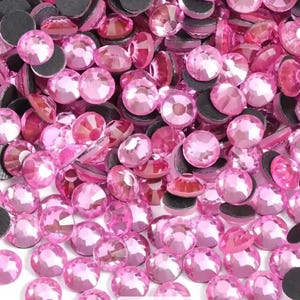 May include: A collection of numerous small, round, faceted pink rhinestones. Each individual stone features a flat, circular top with multiple reflective facets and a dark, opaque circular base, creating a dense, uniform pile of pink gems.