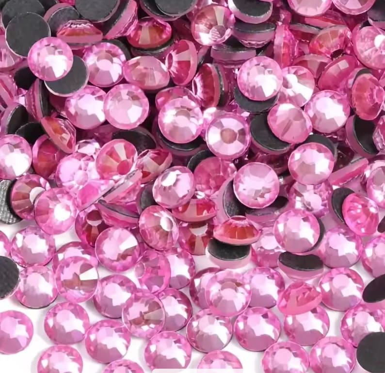 May include: A collection of numerous small, round, faceted pink rhinestones. Each individual stone features a flat, circular top with multiple reflective facets and a dark, opaque circular base, creating a dense, uniform pile of pink gems.