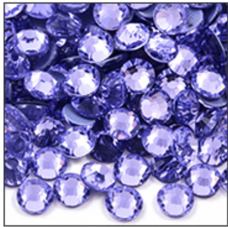 May include: A close-up shot of a pile of round, faceted, light purple rhinestones. The gems are scattered, with varying degrees of light reflection, creating a sparkling effect. The rhinestones are a consistent size and shape.