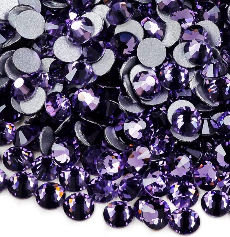 May include: Close-up of a pile of purple rhinestones with silver backing. The faceted crystals are in various sizes and shapes, reflecting light. These decorative embellishments are suitable for crafts and jewelry making.