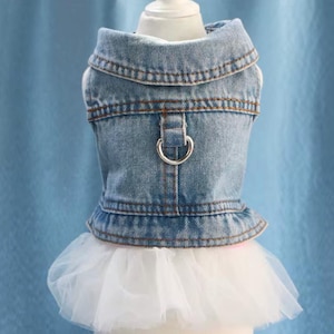 May include: A light blue denim dog vest with a white tulle skirt. The vest has a collar, a metal D-ring, and brown stitching. The tulle skirt adds a touch of elegance to the dog outfit.