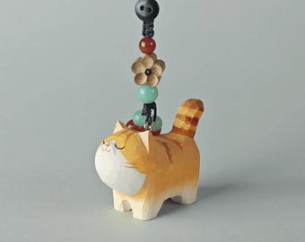 May include: A hand-carved wooden orange tabby cat figurine charm with a white chest and face. The cat is attached to a black braided cord featuring a black button, a red bead, a carved wooden flower, and two light green beads as decorative accents.