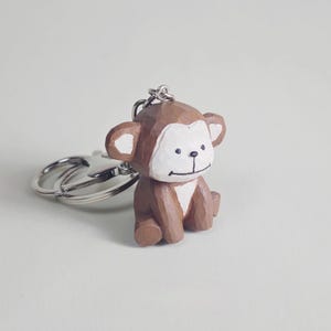 Könnte beinhalten: Ein braun-weißer Affen-Schlüsselanhänger. Der Affe ist aus Holz geschnitzt und hat ein lächelndes Gesicht. Der Schlüsselanhänger hat einen silbernen Metallring und -verschluss.