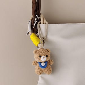 May include: A crocheted teddy bear keychain with a blue bib and white flower detail. The bear is attached to a white and yellow rope, a silver carabiner, and a brown leather strap. The keychain is hanging from a white bag.