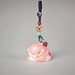 Puede incluir: Una figura de zorro de madera tallada en tono rosa claro, con detalles florales en blanco y rojo. El zorro está en posición de reposo y se sujeta a un cordón trenzado negro decorado con un botón negro, una flor de madera y varias cuentas.