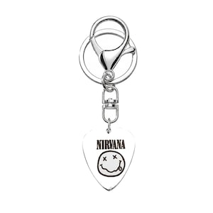 May include: A silver-toned metal keychain featuring a heart-shaped guitar pick charm. The charm displays the black text NIRVANA above a black line-drawn smiley face graphic. The keychain includes a circular ring and a lobster claw clasp.