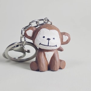 May include: A small, hand-carved wooden monkey keyring featuring a brown and white painted finish. The monkey has a white face with a simple black line for a mouth and two small black dots for eyes. It is attached to a silver-toned metal keyring.