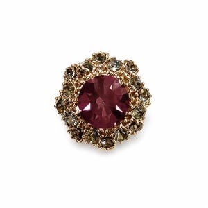 May include: A circular brooch featuring a large, faceted, deep magenta centre stone surrounded by a halo of smaller, dark grey, faceted stones set in a gold-toned metal frame with a textured, scalloped edge design.