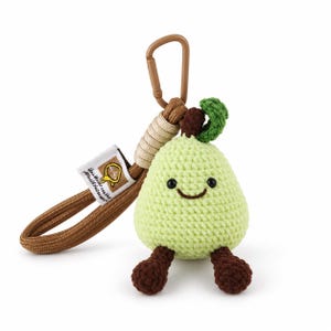 May include: A light green crocheted pear keyring with a smiling face, dark brown legs, a brown stem, and a green leaf. It features a tan paracord loop attached to a brown plastic carabiner and a white fabric tag with a yellow logo and text.