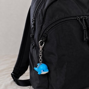 Puede incluir: Una mochila negra con un llavero de mosquetón de metal plateado sujeto a la cremallera frontal. El llavero presenta un pequeño dije de delfín azul brillante con el vientre blanco, acabado liso y pequeños detalles rosados en las mejillas.