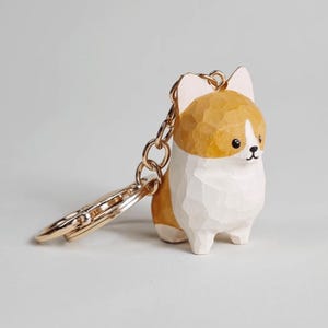 May include: A hand-carved wooden keychain featuring a small dog with a tan and white coat. The figure has a faceted texture, black painted eyes, and a nose. A gold-toned metal chain and ring are attached to the top of the wooden dog figure.