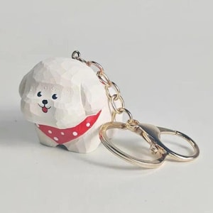 May include: A white carved wooden dog keychain featuring a textured surface, black eyes, a small red tongue, and a red bandana with white polka dots. The dog is attached to a silver-colored metal split ring and a lobster claw clasp connector.