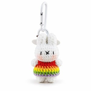 May include: A white crocheted rabbit keyring featuring a dress with horizontal stripes in red, orange, yellow, green, blue, and purple. The rabbit has black embroidered eyes and a cross-shaped mouth, pink cheeks, and is attached to a white carabiner.