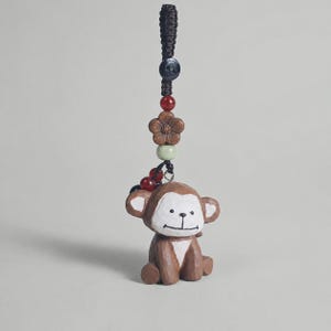 Puede incluir: Un dije de madera tallada con forma de mono en color marrón y blanco, colgado de un cordón trenzado negro. El dije incluye un pequeño botón negro, una cuenta roja, una cuenta marrón con forma de flor y una cuenta verde claro sobre el mono.