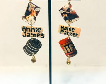 May include: A pair of novelty earrings featuring various pop culture elements. One earring has a picture of a woman, the name "Annie James", and an Oreo cookie. The other has a picture of a man, the name "Hallie Parker", and a Jif peanut butter jar.