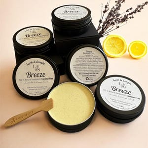 May include: An assortment of black metal tins of Lush & Simple Breeze natural deodorant, with a light yellow balm in an open tin. The labels read "100% Natural Deodorant / Coconut-Free" and "Lavender & Lemon Scent". A small wooden spatula and lemon slices are also present.
