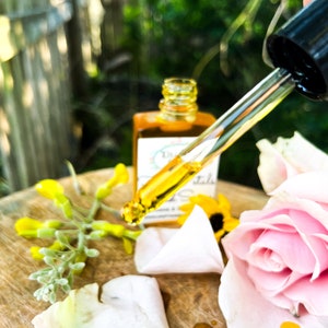 K&ouml;nnte beinhalten: Eine Tropfflasche mit &auml;therischem &Ouml;l mit einer gelben Fl&uuml;ssigkeit, einer rosa Rose und wei&szlig;en Rosenbl&auml;ttern auf einer Holzoberfl&auml;che. Das Etikett der Flasche lautet "Dr. Botanicals Rose & Geranium Oil".