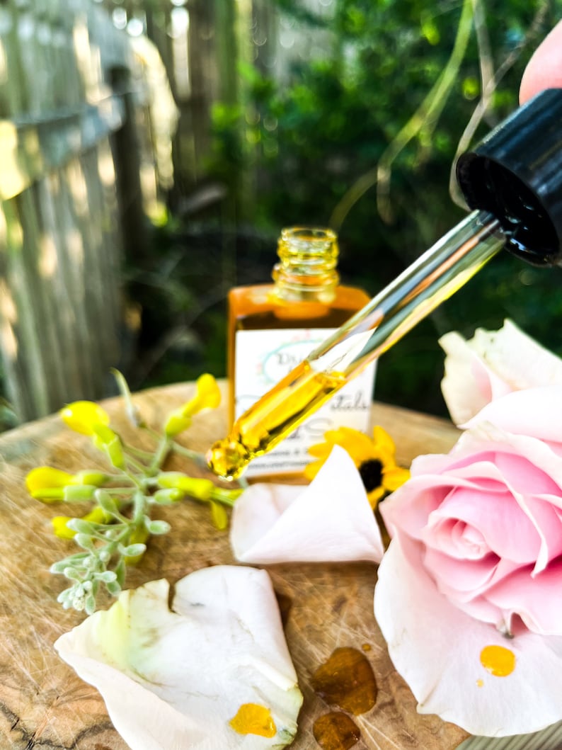 K&ouml;nnte beinhalten: Eine Tropfflasche mit &auml;therischem &Ouml;l mit einer gelben Fl&uuml;ssigkeit, einer rosa Rose und wei&szlig;en Rosenbl&auml;ttern auf einer Holzoberfl&auml;che. Das Etikett der Flasche lautet "Dr. Botanicals Rose & Geranium Oil".