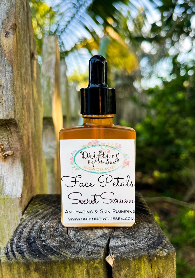 K&ouml;nnte beinhalten: Eine Glasflasche mit Gesichts-Serum und Tropfer. Die Flasche ist mit "Drifting by the Sea Organic Skincare Face Petals Secret Serum Anti-Aging & Skin Plumping WWW.DRIFTINGBYTHESEA.COM" beschriftet.