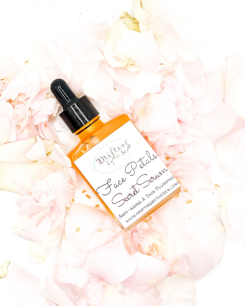 K&ouml;nnte beinhalten: Eine durchsichtige Glasflasche mit schwarzem Tropfer, gef&uuml;llt mit einer gelben Fl&uuml;ssigkeit. Die Flasche ist mit "Drifting by the Sea Face Petals Secret Serum Anti-Aging & Skin Plumping www.driftingbythesea.com" beschriftet. Die Flasche ist von rosa Rosenbl&auml;ttern umgeben.