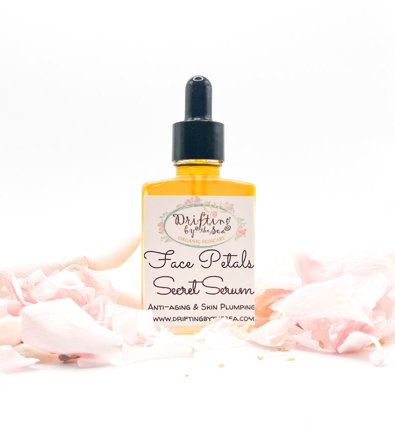 K&ouml;nnte beinhalten: Eine Glasflasche mit Gesichts-Serum und Tropfer. Die Flasche ist mit "Drifting by the Sea" und "Face Petals Secret Serum" beschriftet. Das Etikett tr&auml;gt au&szlig;erdem die Aufschrift "Anti-Aging & Skin Plumping" und "www.driftingbythesea.com". Die Flasche ist von rosa Rosenbl&auml;ttern umgeben.