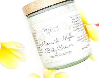 Może przedstawiać: Szklany słoik z bambusową pokrywką wypełniony białym kremem do ciała. Słoik ma etykietę z napisem "Drifting by the Sea Mermaid Mylk Body Cream Beach Cottage". Etykieta zawiera również tekst "THE RITUAL skin morning Excellent for damaged, dry mylky body smooth and h dry skin Use on dry a elbows, and c could uses www.drifti"