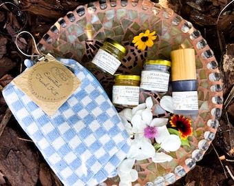 May include: A blue and white checkered travel kit with a tag that reads "Essential Travel Kit" and four small jars of face care products, including a face balm, face cleanser, cleansing oil, and lavender serum. The products are arranged on a mosaic tile surface with flowers.