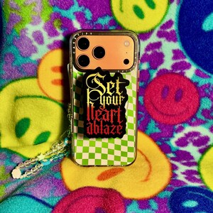 May include: A clear phone case with a green and white checkered pattern and a sticker that says "Set your heart ablaze" in black and red. The phone case has an orange camera section and a clear beaded wrist strap. The background is a colorful fabric with smiley faces.