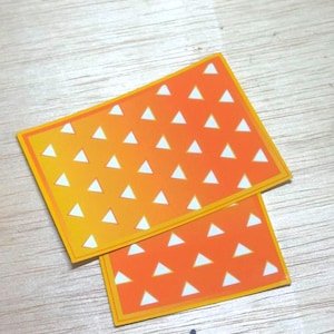 May include: Two rectangular stickers with an orange and yellow gradient background. Each sticker features a pattern of white triangles and a yellow border. The stickers are overlapping on a light wood surface.