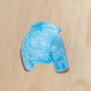 May include: A light blue, puffy winter jacket with white polka dots. The jacket is a sticker or cutout, and is set against a light wood grain background. The jacket has a collar and two sleeves.