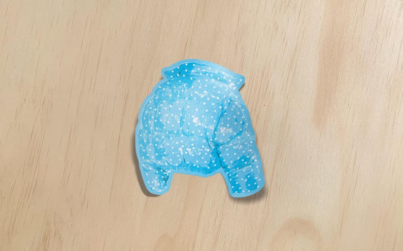May include: A light blue, puffy winter jacket with white polka dots. The jacket is a sticker or cutout, and is set against a light wood grain background. The jacket has a collar and two sleeves.