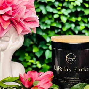 May include: A black candle with a wooden lid, labeled "LaBella's Fruition" from the G7 Collection. The candle is 4 oz (113 g) and made with coconut apricot creme wax. Pink flowers and a white ceramic bust are in the foreground.