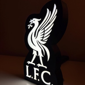 Può includere: Un'insegna luminosa in bianco e nero con l'emblema del Liver Bird e le lettere "L.F.C.". L'insegna è un design tridimensionale, con il Liver Bird in bianco e lo sfondo in nero. Le lettere "L.F.C." sono anch'esse bianche.